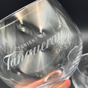 May include: Clear, stemmed gin glass with the words "TONIGHT WE Tanqueray" etched in white script. The glass is angled, with a second glass visible in the background. The image is well-lit, highlighting the glass's clarity.