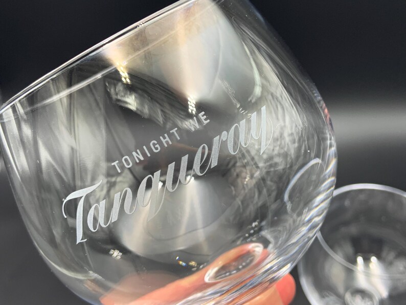 May include: Clear, stemmed gin glass with the words "TONIGHT WE Tanqueray" etched in white script. The glass is angled, with a second glass visible in the background. The image is well-lit, highlighting the glass's clarity.