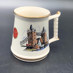 Könnte beinhalten: Cremefarbene Keramiktasse mit goldfarbenem Rand und Henkel, die die Tower Bridge in London mit einem roten Bus zeigt. Die Tasse hat einen roten Aufkleber. Auf der Tasse steht "Tower Bridge".