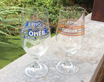 May include: Two clear beer glasses with the word "OMER" in blue and white lettering. The glasses have a heart-shaped design etched on the lower portion. One glass has blue accents, the other has orange accents.