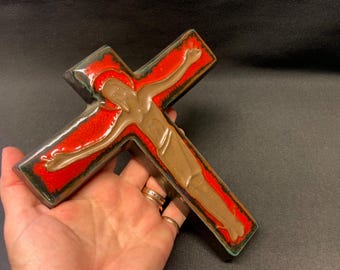 May include: A ceramic cross with a figure of Jesus. The cross is primarily red with a brown figure and a dark green border. The figure has outstretched arms. The cross is held in a hand.