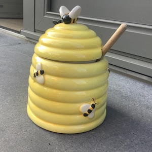 Può includere: Un vasetto di miele in ceramica gialla a forma di alveare, con un cucchiaio di legno. Il vasetto ha un coperchio con un'ape sopra e api decorative sui lati. Il vasetto è alto circa 15 cm.