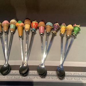 Könnte beinhalten: Eine Sammlung von Eiscremelöffeln mit bunten Griffen in Form von Eiswaffeln. Die Löffel haben lange, schlanke, silberfarbene Stiele und abgerundete Schalen. Die Eiswaffelgriffe sind in verschiedenen Farben gehalten.