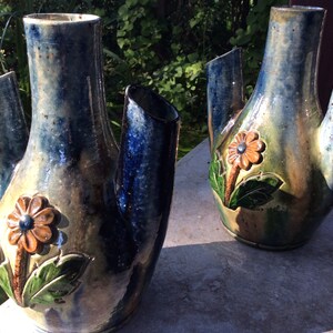 Könnte beinhalten: Zwei blaue und braune Keramikvasen mit jeweils drei Ausgüssen. Die Vasen haben ein florales Design mit orangefarbenen Blumen und grünen Blättern.