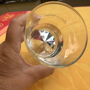 May include: A clear, faceted-bottom glass. The glass is held at an angle, revealing the interior design. The rim of the glass has a slight curve. The glass is on a yellow surface.