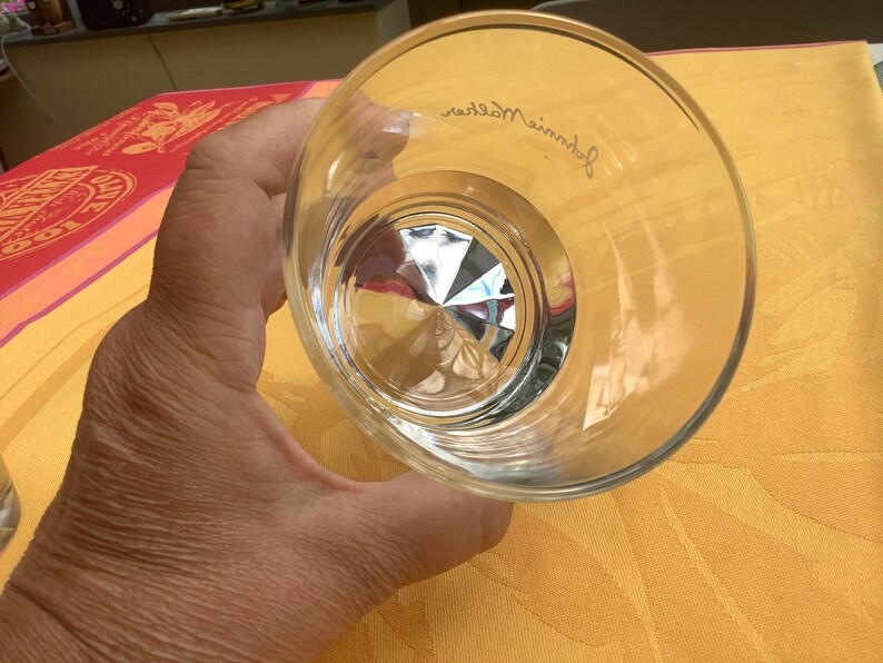 May include: A clear, faceted-bottom glass. The glass is held at an angle, revealing the interior design. The rim of the glass has a slight curve. The glass is on a yellow surface.