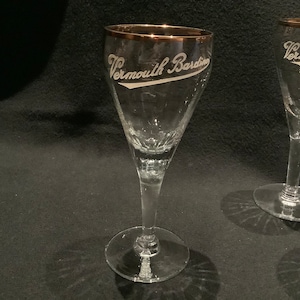 Op de afbeelding: Twee heldere glazen op voet met een gouden rand aan de bovenkant. Elk glas is voorzien van de witte geëtste tekst Vermouth Bardino aan de zijkant. De glazen hebben een taps toelopende kelkvorm, een slanke steel en een ronde voet voor dranken.