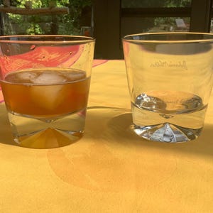 May include: Two clear, lowball glasses on a yellow tablecloth. One glass contains a brown liquid with ice cubes. The other glass is empty, with the name "Johnnie Walker" etched on the side. The glasses have a geometric base.