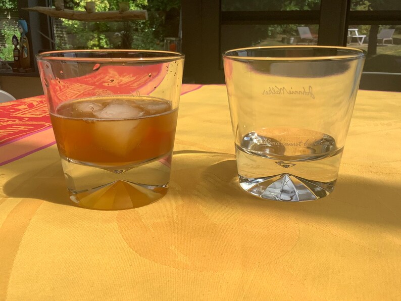 May include: Two clear, lowball glasses on a yellow tablecloth. One glass contains a brown liquid with ice cubes. The other glass is empty, with the name "Johnnie Walker" etched on the side. The glasses have a geometric base.