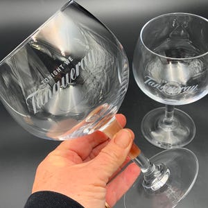 May include: Two clear, stemmed gin glasses with the Tanqueray logo and the words "TONIGHT WE" etched on the glass. One glass is held by a person. The other glass is in the background. The glasses are on a dark surface.