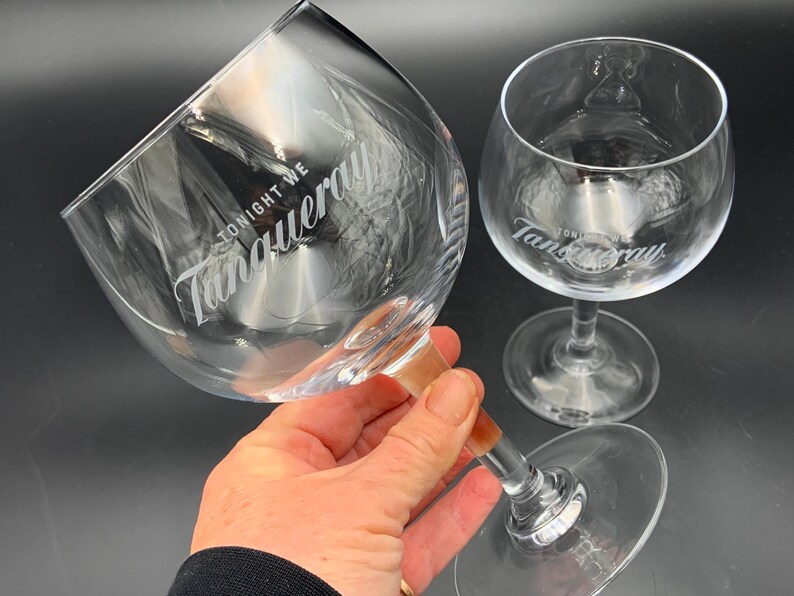 May include: Two clear, stemmed gin glasses with the Tanqueray logo and the words "TONIGHT WE" etched on the glass. One glass is held by a person. The other glass is in the background. The glasses are on a dark surface.