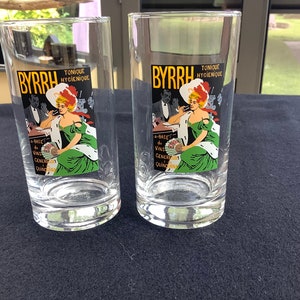 May include: Two clear glass tumblers with a vintage advertisement for Byrrh tonic wine. The advertisement features a woman in a green dress and a white hat, holding a glass of wine. The text on the advertisement reads "Byrrh, Tonique Hygiénique, A base de vins genereaux du Quina-quina."