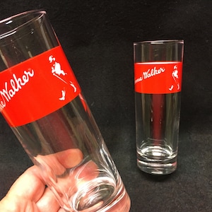 May include: Two clear glass tumblers with a red band around the middle. The band has the words "Johnnie Walker" printed in white. The glass on the left has a white silhouette of a man walking.