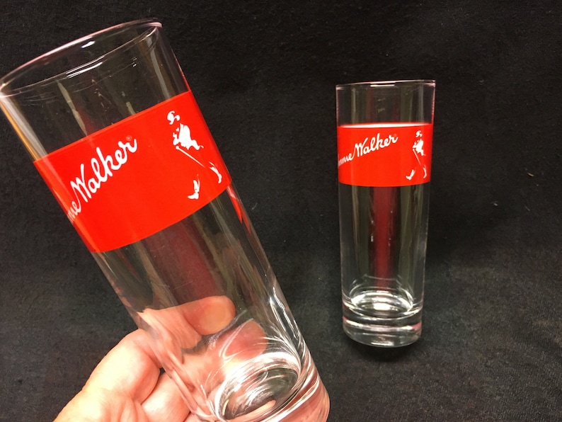 May include: Two clear glass tumblers with a red band around the middle. The band has the words "Johnnie Walker" printed in white. The glass on the left has a white silhouette of a man walking.
