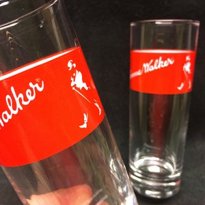 May include: Two clear glass tumblers with a red band around the middle. The band has the words "Johnnie Walker" in white lettering and a white silhouette of a man walking.