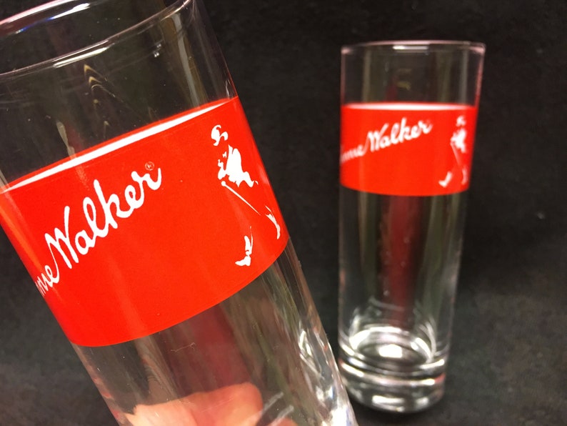May include: Two clear glass tumblers with a red band around the middle. The band has the words "Johnnie Walker" in white lettering and a white silhouette of a man walking.