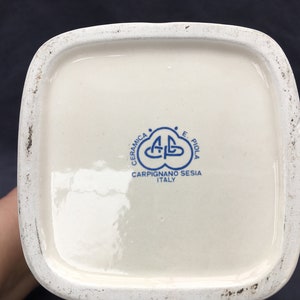 Pode incluir: Vista inferior de um objeto de cer&acirc;mica branca com formato quadrado. Apresenta um logotipo azul que diz "CERAMICA E. PIOLA CARPIGNANO SESIA ITALY". As bordas mostram sinais de desgaste.