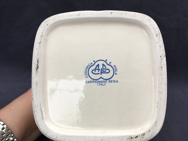 Pode incluir: Vista inferior de um objeto de cer&acirc;mica branca com formato quadrado. Apresenta um logotipo azul que diz "CERAMICA E. PIOLA CARPIGNANO SESIA ITALY". As bordas mostram sinais de desgaste.