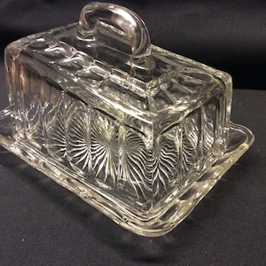 Op de afbeelding: Transparante glazen botervloot met een rechthoekige vorm en een handvat op het deksel. De schaal heeft een decoratief, gestructureerd patroon op de zijkanten en het deksel en staat op een bijpassende basis. Een klassiek keukenartikel.