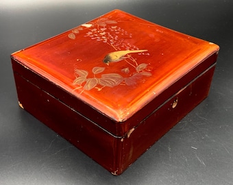 Può includere: Una scatola rettangolare in legno laccato con finitura rossa lucida. Il coperchio presenta un disegno di uccello e fiori dipinto in oro e marrone. La scatola ha un piccolo buco della serratura sul davanti.