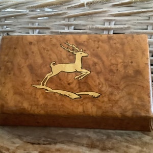 Peut inclure: Boîte rectangulaire en bois avec une finition marron clair et un motif de cerf doré en train de sauter. La boîte présente un motif décoratif en loupe de bois, probablement pour le rangement ou l'exposition.