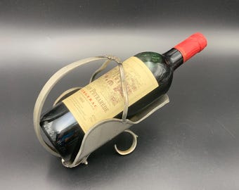 May include: A silver-colored wine bottle holder cradles a bottle of red wine with a red foil top. The bottle has a yellow label with text. The holder has a curved handle and a curved base.