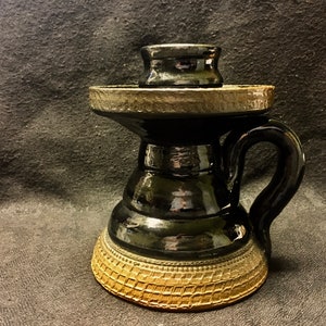 Peut inclure: Une tasse en céramique noire avec une anse et un couvercle. La tasse présente un design texturé et étagé et une base décorative avec un motif tissé. La poignée est incurvée et le couvercle est posé dessus. La tasse mesure environ 15 cm de haut.