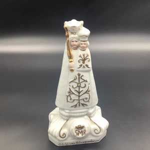 Może przedstawiać: Biała figurka z porcelany przedstawiająca Matkę Boską z Scherpenheuvel, katolicką postać religijną, z koroną ze złota i ozdobnymi złotymi detalami. Figurka stoi na podstawie z napisem "O.L.V. van Scherpenheuvel" i "SMR".