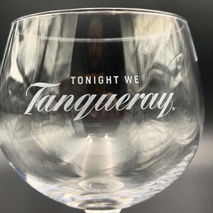 May include: Clear, stemmed gin glass with the words "TONIGHT WE Tanqueray" in white script. The glass has a rounded bowl and a textured base. The image is a close-up, with a dark background.