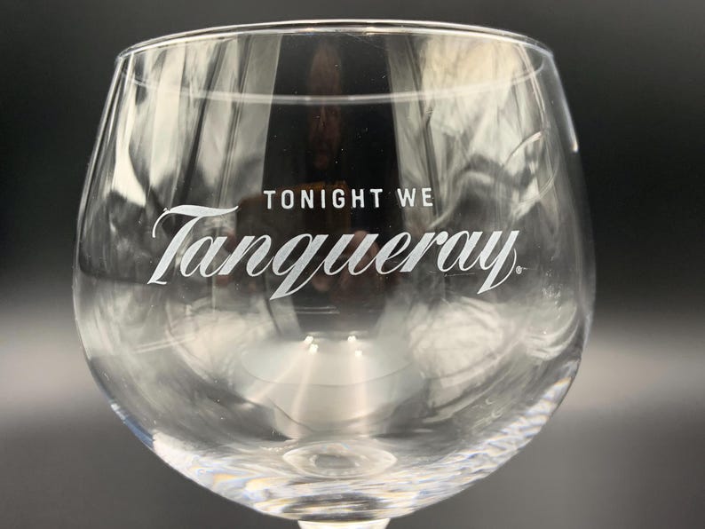 May include: Clear, stemmed gin glass with the words "TONIGHT WE Tanqueray" in white script. The glass has a rounded bowl and a textured base. The image is a close-up, with a dark background.