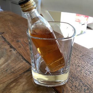 May include: A miniature bottle of amber-colored liquor sits inside a clear glass tumbler with a walking man logo on the bottom.