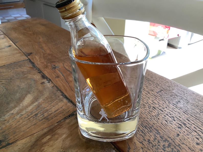 May include: A miniature bottle of amber-colored liquor sits inside a clear glass tumbler with a walking man logo on the bottom.