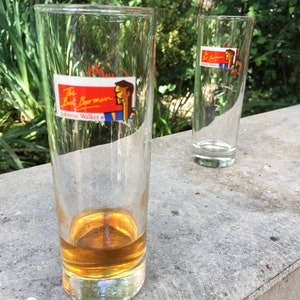 May include: Two tall, clear glass tumblers. One tumbler is filled with a golden brown liquid. Both tumblers have a red, white, and blue sticker with the text "The Bart Barman Johnnie Walker" and a cartoon image of a man.