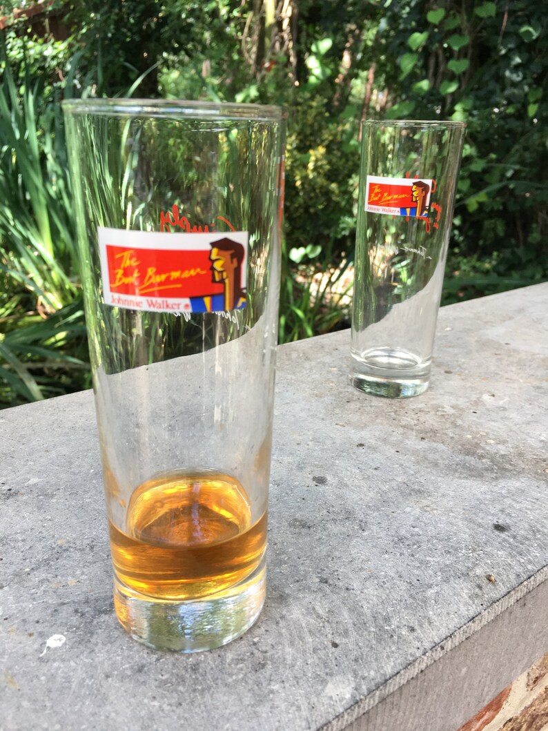 May include: Two tall, clear glass tumblers. One tumbler is filled with a golden brown liquid. Both tumblers have a red, white, and blue sticker with the text "The Bart Barman Johnnie Walker" and a cartoon image of a man.