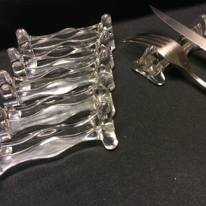 Pode incluir: Conjunto de seis apoios para faca de vidro transparente com um design ondulado. Os apoios para faca estão empilhados uns sobre os outros.