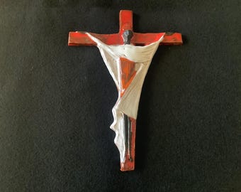 May include: A red wooden cross with a white and red figure of a person on it. The figure is wearing a white robe and has a black face.