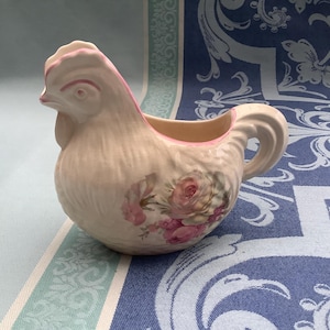 Może przedstawiać: Ceramiczny dzbanek w kształcie kury z różowym grzebieniem i akcentami. Dzbanek ma kwiatowy wzór z różowymi różami. Uchwyt jest zakrzywiony, a dzbanek jest ustawiony na niebiesko-białym tle.