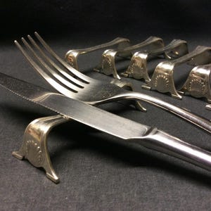 Può includere: Coltello e forchetta in metallo argentato su un supporto in metallo argentato. Il supporto è progettato per tenere un coltello e una forchetta in posizione verticale. Ci sono diversi altri supporti sullo sfondo.