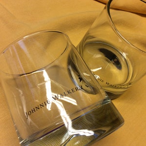 May include: Two clear glass tumblers with the words "JOHNNIE WALKER" printed on the side. The glasses are sitting on a yellow surface.