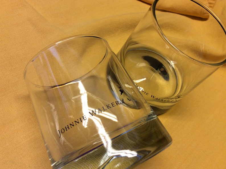 May include: Two clear glass tumblers with the words "JOHNNIE WALKER" printed on the side. The glasses are sitting on a yellow surface.