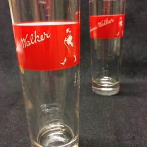 May include: Two clear glass tumblers with a red band and white "Johnnie Walker" logo. The logo includes a stick figure of a man walking with a cane.