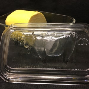 May include: A clear glass butter dish with a lid featuring an embossed cow design. The dish contains a block of yellow butter. The lid has a rectangular shape with rounded corners. The butter dish is a kitchenware item.