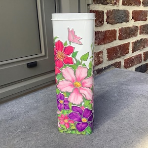 Könnte beinhalten: Eine weiße rechteckige Dose mit einem Blumendesign mit rosa, roten und lila Blumen mit grünen Blättern. Die Blumen sind handbemalt auf der Dose. Die Dose steht auf einer grauen Oberfläche.