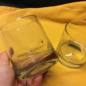 May include: Two clear glass tumblers with the Johnnie Walker logo printed on the bottom. The tumblers are on a yellow surface.