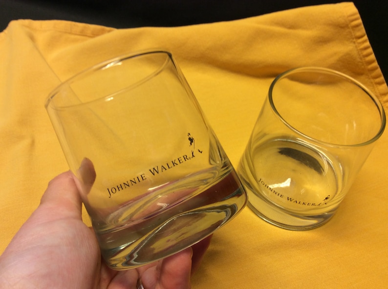 May include: Two clear glass tumblers with the Johnnie Walker logo printed on the bottom. The tumblers are on a yellow surface.