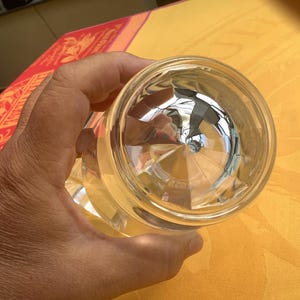 May include: Clear, faceted glass with a geometric design. The glass is held in a hand, with the bottom of the glass facing the camera. The glass reflects the surrounding environment, creating a mirrored effect. The glass is likely a drinking glass or decorative item.
