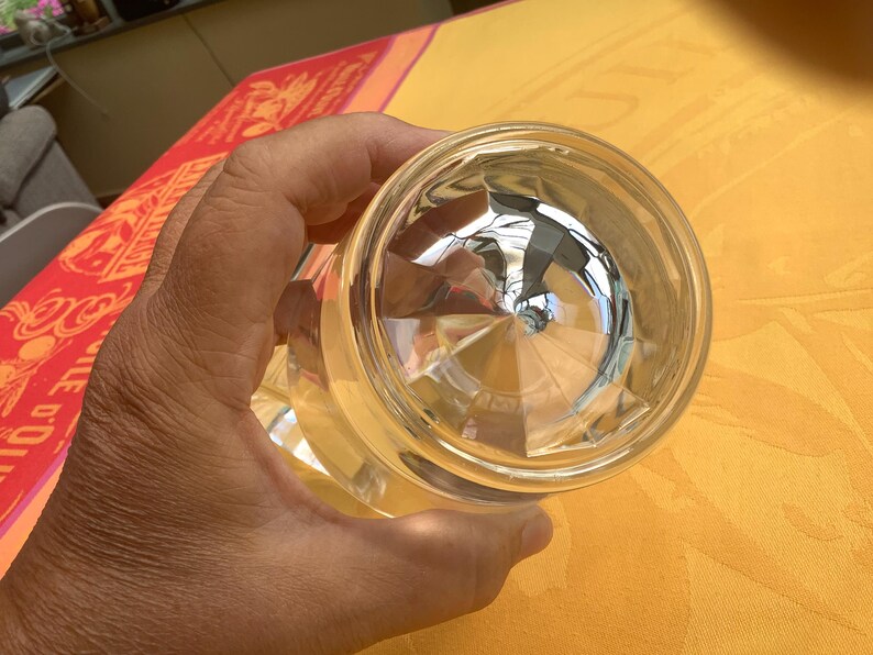 May include: Clear, faceted glass with a geometric design. The glass is held in a hand, with the bottom of the glass facing the camera. The glass reflects the surrounding environment, creating a mirrored effect. The glass is likely a drinking glass or decorative item.