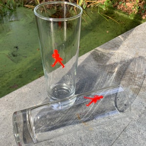 Op de afbeelding: Twee heldere glazen tumblers met een rood lopend figuurlogo. De glazen staan op een grijs oppervlak.