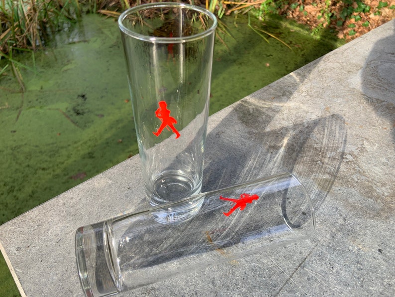 Op de afbeelding: Twee heldere glazen tumblers met een rood lopend figuurlogo. De glazen staan op een grijs oppervlak.