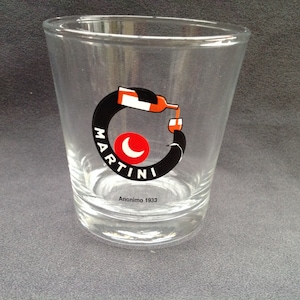 Op de afbeelding: Een helder glazen tumbler met een zwart-wit Martini-logo. Het logo heeft een rode halve maan en een fles die in een glas giet. De tekst "Anonimo 1933" staat gedrukt op de bodem van het glas.